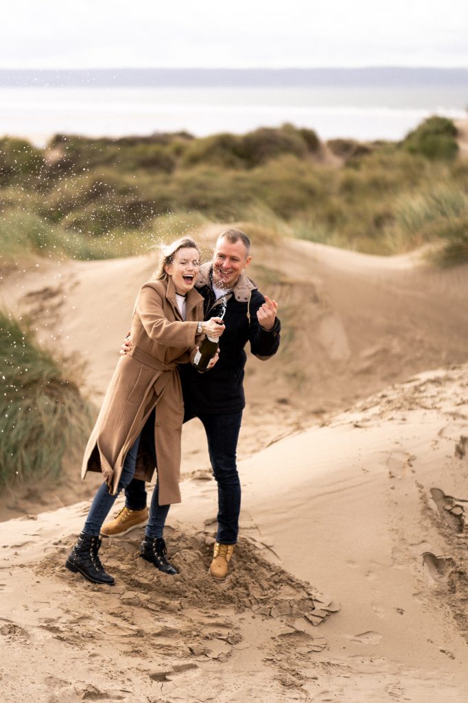 Couple on beach celebrating, popping champagne bottle with spray.